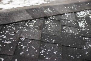Hailstones scattered across dark asphalt roof shingles after a hailstorm
